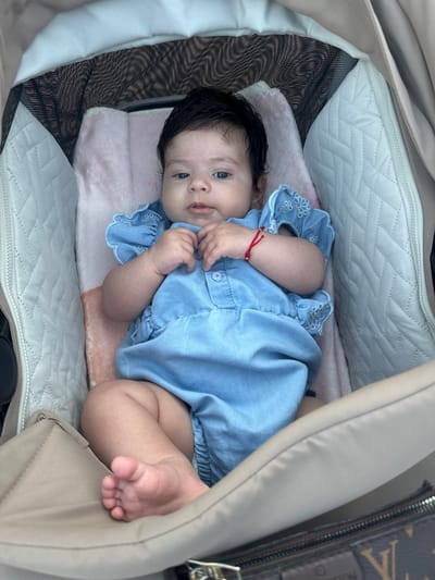 A baby girl is lying in a stroller. She is wearing a blue romper and has a red string bracelet on her left wrist. The stroller has a light-colored interior with quilted padding and a mesh canopy. The background is blurred, but suggests an outdoor setting. The location is the city of Svishtov, Bulgaria.