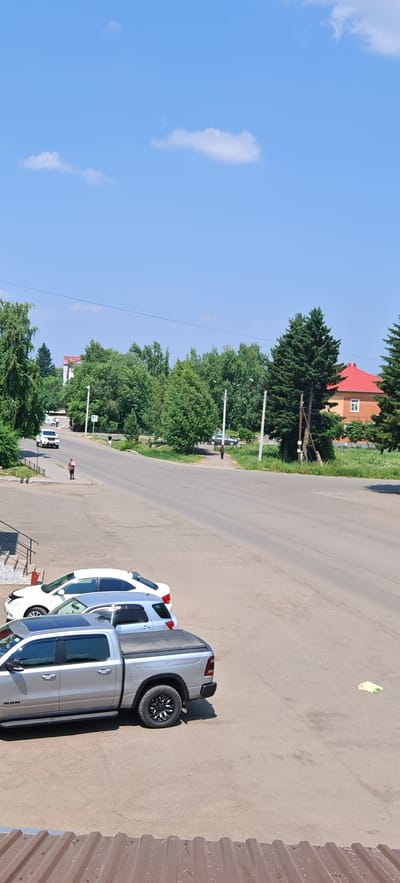 The image depicts a street scene in Altayskoye, Russia, under a clear blue sky with a few scattered clouds. In the foreground, a silver pickup truck with a black tonneau cover is parked on a dirt lot. To its left are a silver SUV and a white sedan. Further up the street, a white SUV is driving away from the camera, and to the left of the road, a person in a pink shirt is walking away. Across the street, there are several trees, a building with a red roof, and another person standing. Telephone poles are visible along the side of the road. The overall scene suggests a quiet, sunny day in a small town.