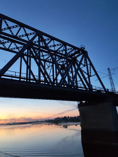 The image depicts a steel truss bridge, silhouetted against a gradient sky transitioning from orange near the horizon to deep blue above. The bridge supports a railway track and has a concrete support structure visible on the right. Below the bridge, a calm body of water reflects the colors of the sky and the faint lights of the distant shoreline, which is obscured by a band of mist. The location is identified as Tarkanovo, Russia.
