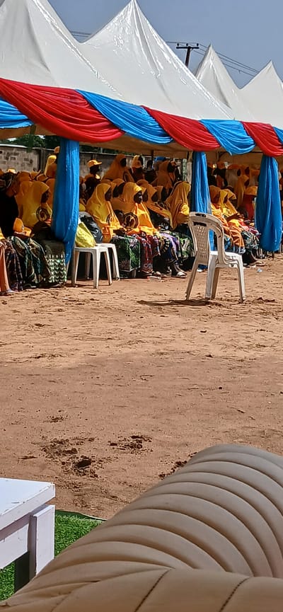 In Rigachikun, Nigeria, under a white tent adorned with red and blue fabric, a group of people, predominantly women, are seated. They are dressed in yellow attire and headscarves, with some wearing matching yellow caps. The ground beneath them is sandy. In the foreground, a white plastic chair stands empty. To the left, a white table is partially visible, with a patch of green grass behind it. The scene suggests an outdoor gathering or event.