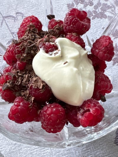 A close-up, overhead shot showcases a serving of raspberries in a clear glass bowl. The bowl is filled with vibrant red raspberries, some of which are partially obscured by dark chocolate shavings. A dollop of a creamy white substance, possibly yogurt or whipped cream, sits in the center of the raspberries and chocolate. The background is a neutral gray and white patterned fabric. The lighting appears to be natural and diffused, suggesting it might be daytime. There is no visible text in the image. The scene appears to be a simple, appealing dessert or snack preparation.