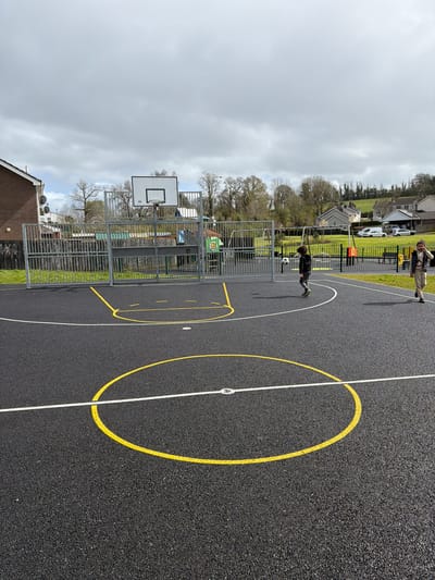 A view of an outdoor basketball court with a black asphalt surface marked with yellow and white lines. A basketball hoop is positioned behind a gray metal fence. Two people are in the background, one dribbling a soccer ball. The background shows a residential area under an overcast sky. Based on the general architectural style and the presence of a basketball court, it is possible that the scene is in Enniskillen, United Kingdom.