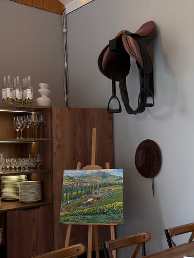The image depicts an indoor scene, likely a room in Dsegh, Armenia, with a rustic and artistic ambiance. Dominating the upper wall is a brown leather horse saddle mounted on a black bracket, with stirrups hanging down. To the left, on a shelf, are several clear glass bottles with white labels, and a white, tiered decorative vase. Below this shelf, within a dark wood cabinet, are rows of clear wine glasses on two shelves, and stacked plates on a lower shelf. To the right of the cabinet, an easel holds a landscape painting of a rural scene with sunflowers in the foreground and rolling hills in the background. Below the saddle, on the wall, hangs a brown leather hat. Two wooden chairs with dark metal frames are partially visible in the lower foreground. The overall lighting suggests an interior space with artificial lighting.
