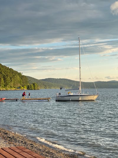 The image depicts a serene lakeside scene in Turgoyak, Russia, under a partly cloudy sky. In the foreground, a wooden dock made of several planks extends into the water. The lake's surface is rippled by gentle waves, with a pebbly shoreline visible on the left.

Several objects are present on the water. A white sailboat with a tall mast and furled sails is moored in the middle ground, its hull bearing the name "GYPSEA" and the registration "AH1371RUS78". To the left of the sailboat, a smaller wooden pier juts out from the shore. On this pier, two figures are standing: one person in a red jacket and yellow boots, and a smaller figure, likely a child, in dark clothing. A red kayak or boat is partially visible moored to the end of this pier.

Further out on the lake, a pontoon-like structure is floating, bearing the Cyrillic inscription "АКВАЛАЙФ" (AQUALIFE). The background is dominated by a lush, green, forested hillside that slopes down towards the water, with rolling hills extending into the distance under the soft, diffused light of the sky. The overall atmosphere is peaceful and idyllic, suggestive of a recreational area on the lake.