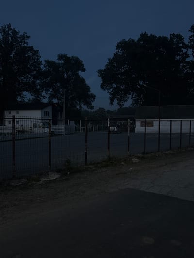 The image depicts a dimly lit outdoor scene, likely taken at dusk or dawn due to the low light and dark blue sky. The orientation of the image is rotated 90 degrees counter-clockwise.

The foreground is dominated by a dark asphalt road on the right, with a strip of dry grass and dirt along its left edge. To the left of the grass is a wire mesh fence, supported by dark posts. Beyond the fence, a building with a white and grey facade is visible, featuring several windows. A large tree with dark foliage partially obscures the building and the sky.

In the background, more trees and structures are faintly visible through the fence, suggesting a semi-rural or suburban setting. The overall atmosphere is quiet and still, with no visible human activity. The lighting suggests a cool temperature, and there are no clear indications of specific weather conditions beyond the low light. The location context provided is Slobozia-Dușca, Moldova. No visible text is present in the image.