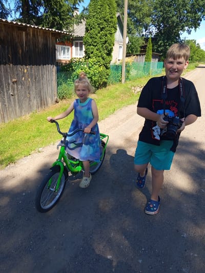 In Ostrovtsy, Russia, a young boy, possibly pre-teen, stands on a dirt path, holding a black Canon digital camera around his neck. He wears a black t-shirt with graphic print and turquoise shorts, along with blue Crocs decorated with colorful charms. He smiles broadly, looking directly at the camera. Beside him, a younger girl, likely a toddler, sits on a bright green bicycle. She wears a blue and green tie-dye dress and cream-colored Crocs. Her hair is tied back in a small bun, and she looks towards the camera with a slight smile. The scene is set outdoors on a sunny day, with lush green grass and trees surrounding the dirt path. A wooden fence and parts of a house are visible in the background. The time of day appears to be midday due to the bright sunlight and distinct shadows. The overall atmosphere is cheerful and relaxed, suggesting a casual outdoor activity. Text visible on the boy's camera strap reads "Canon" and "EOS DIGITAL." The bicycle frame also has the brand name "STELS" visible.