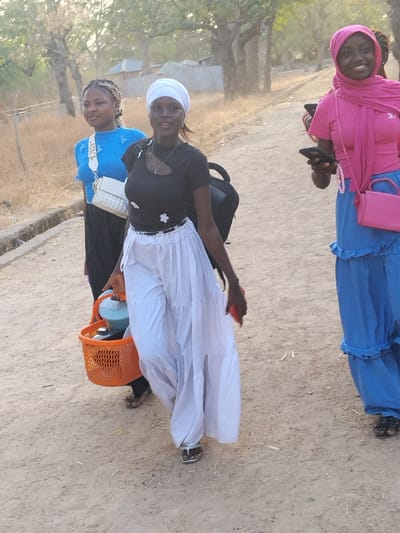 Three young women are walking on an unpaved dirt path in Garun Gawa, Nigeria. In the foreground, a woman wears a black short-sleeved top with shoulder cutouts, white tiered wide-leg pants, white sandals, and a white headwrap. She carries an orange plastic basket containing a light blue thermal pot and other items in her left hand, and a red smartphone in her right hand. A black backpack is visible on her back. She is smiling and looking towards the camera. To her left, slightly behind, a second woman wears a light blue long-sleeved top, dark trousers or a long skirt, and a white cross-body bag with a wide strap. Her hair is styled in braids. She is also smiling. To the right, further back, a third woman wears a pink short-sleeved top, a pink hijab, a blue tiered long skirt, and black sandals. She carries a pink cross-body bag and holds a dark smartphone in her left hand. She is smiling broadly. The environment consists of a dusty, unpaved path with sparse, dry vegetation and several trees with minimal foliage lining the sides. A low concrete structure or drain runs along the left edge of the path. Distant, indistinct structures are visible in the background under natural daylight.