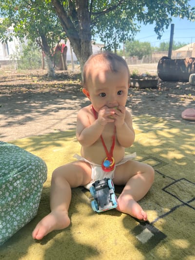 This is a vertical, eye-level, outdoor, full shot of a baby playing on a yellow and green patterned rug. The baby is lying on their back, facing the camera, with their head tilted slightly to the right. They are wearing only a diaper and have a pacifier with a red string around their neck. The baby is holding a small green object in their mouth. A small toy car with blue wheels is placed near their right foot. The rug is on a dirt and gravel ground. In the background, there are trees with green leaves, some fallen leaves on the ground, and what appears to be a fence and some structures. The lighting suggests it is daytime, with dappled sunlight filtering through the trees, creating shadows on the rug. The overall mood appears relaxed and playful, with the baby seemingly engaged with their toy and the object in their mouth. There is no visible text in the image. The location context provided is Айдарлы, Kazakhstan.