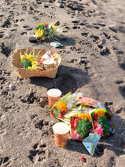 The image depicts a beach setting in Tibubeneng, Indonesia, featuring offerings placed on the sand. A woven basket holds several items, including small wrapped parcels, a bottle of liquid, and a cup labeled "MAY TO COFFEE". Nearby is another arrangement with cups, floral and vegetative arrangements, and a folded banknote. The sand is dark and shows various footprints. The light suggests it is daytime. No people are visible in the shot.