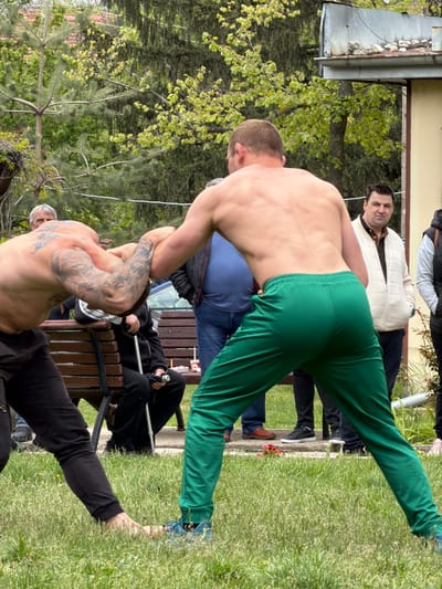 The image depicts an outdoor setting in Dulevo, Bulgaria, with at least 8 people present. Two shirtless men are engaged in arm wrestling, with one man wearing green pants and the other wearing black pants and displaying prominent tattoos on his left arm. Other individuals are watching the arm wrestling match, some seated on a bench. The environment includes grassy areas, trees, and a building.