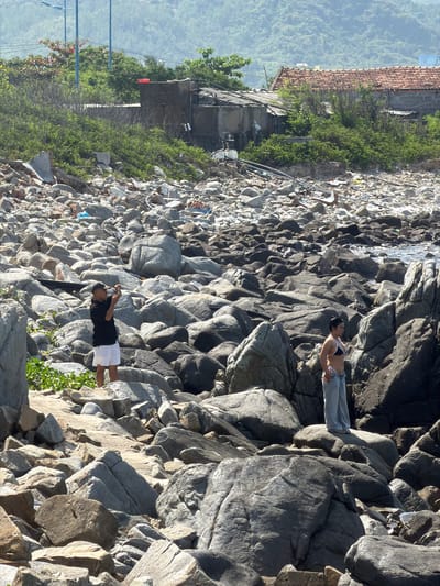 The image depicts a rocky shoreline in Vĩnh Lương, Vietnam. In the foreground, a man wearing a black t-shirt and white shorts stands on a rock, holding a phone to take a photo. To the right, a woman in a bikini top and light-colored pants stands on another rock, facing away from the camera. The shoreline is covered in various sizes of grey and brown rocks, with some green foliage growing among them. In the background, there are some small buildings with tiled roofs and a lush green hillside. The sea is visible in the distance, with calm water reflecting the sunlight.