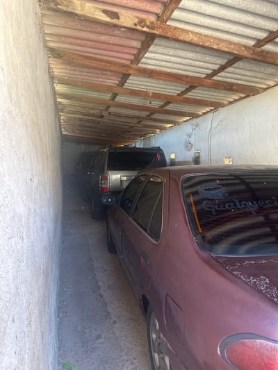 The image shows an outdoor covered parking area. Two vehicles are parked closely together. The vehicle in the foreground is a maroon sedan, and the vehicle behind it appears to be a dark-colored SUV. The parking area has a rough, uneven concrete floor and is enclosed by rough, light-colored concrete walls. The roof is constructed of corrugated metal sheeting supported by wooden beams. There is graffiti or writing on the rear window of the maroon sedan. The scene is lit by natural light filtering into the covered area. The location is identified as being in El Maco, Venezuela.