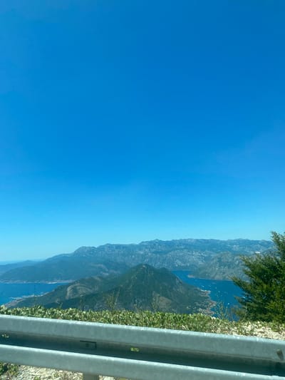 The image captures a panoramic view of a mountainous coastal region under a clear blue sky. In the foreground, a metal guardrail runs horizontally across the bottom of the frame, with green foliage growing just beyond it. The middle ground reveals a dramatic landscape of dark green, tree-covered mountains sloping down towards a bright blue body of water. The water is dotted with numerous small white specks, likely boats. Further in the background, a range of lighter-colored, rugged mountains stretches across the horizon. The scene is a scenic vista, likely from a high vantage point, showcasing the natural beauty of the area. While the prompt specifically mentions Žanjev Do, Montenegro, the image itself does not contain explicit landmarks or identifiers that confirm this precise location without further context. However, the geography is consistent with the mountainous coastline of Montenegro. There are no people, animals, or discernible actions occurring in the image.