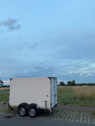 A white, box trailer with two axles and dual wheels is parked on a permeable pavement surface adjacent to a paved road. The trailer is positioned in front of a grassy field with tall, dry grass and some scattered trees. In the background, there are more trees, some industrial structures, and a building with a dark roof on the left side of the frame. The sky is overcast with soft blue and grey clouds, indicating either early morning or late evening. The scene appears to be located in an undeveloped or semi-developed area of Bönnigheim, Germany, possibly on the outskirts of the town, given the presence of open fields and a view of distant industrial elements. There are no people or animals visible in the image.