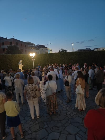 The image depicts a group of people gathered in an outdoor courtyard at dusk in Međugorje, Bosnia and Herzegovina. In the foreground, several individuals are seen from behind, with a young boy in blue shorts and a light yellow shirt standing in the center. The ground is paved with irregular stone tiles. Behind them, a crowd of adults stands facing a white statue of a woman, likely the Virgin Mary, illuminated by a bright lamp post with two globes. A low, manicured hedge forms a backdrop behind the statue and the lamp post. In the distance, buildings are visible against the twilight sky. The overall atmosphere is one of a religious gathering or pilgrimage.