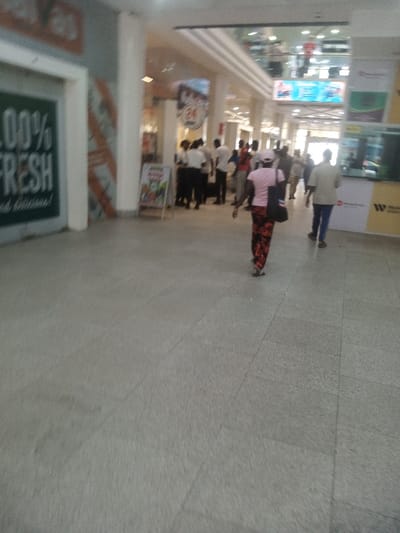The image depicts an indoor commercial corridor, likely within a shopping mall or large building, located in Majengo ward, Kenya. The floor is covered with light grey, rectangular tiles. On the left, a storefront features a dark green wall with "100% FRESH" in white text and diagonal orange and white striped accents. An A-frame sign with colorful imagery is positioned on the floor. Behind this sign, a group of approximately 8-10 individuals, many in white shirts and dark trousers, are standing near a shop entrance marked by a "24" sign on a red background.

In the foreground, a person walks away from the camera, wearing a pink short-sleeved top, multi-colored patterned trousers (red, black, white), and light-colored sandals, carrying a dark shoulder bag. Further into the mid-ground on the right, another person walks away, dressed in a light-colored shirt and dark trousers. The background shows a well-lit space with multiple white pillars and reflections from overhead lighting on the ceiling. On the right wall, there are visible logos for "Western Union" and "moneyGram" on a yellow and white panel. Numerous blurred figures indicate additional foot traffic throughout the corridor.