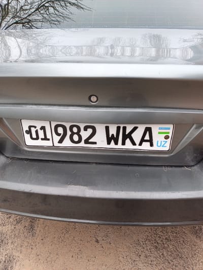 The image displays the rear of a grey car, focusing on the license plate. The license plate reads "01 982 WKA", with "UZ" (Uzbekistan) indicated on the right. The plate is white with black lettering, and features the Uzbekistan flag emblem. A circular sensor or button is located above the license plate. The bumper is also grey and shows some minor signs of wear. A reflection of trees can be seen in the car's rear window.