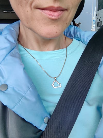 The image is a close-up shot of a person's neck and chest area, focusing on the lower part of their face and their clothing. The person is wearing a light blue crewneck sweatshirt and a light blue puffer jacket. A thin gold chain necklace with a pendant shaped like a map of Belarus is visible on the sweatshirt. A black seatbelt is diagonally across the chest, obscuring part of the body and clothing. The background is out of focus but appears to be an outdoor setting, possibly a street or a parking lot, with hints of a building and a streetlamp. The lighting suggests it is daytime. The person's skin is fair with visible freckles and minor blemishes. The scene appears to be taken from inside a vehicle, given the presence of the seatbelt and the focus on the individual. The provided location "Фаніпальскі сельскі Савет, Belarus" is context for the map pendant, not a detail directly visible in the image itself.