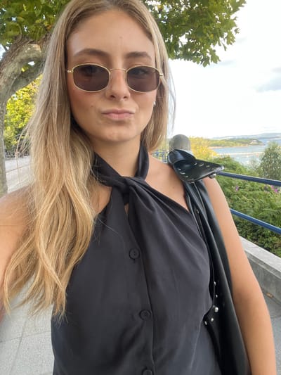 The image displays a blonde woman, likely a young adult, captured in a close-up, outdoor portrait, presumably a selfie, overlooking Santander Bay in Santander, Spain. She is positioned slightly to the left of the frame's center. The woman wears gold-framed rectangular sunglasses with dark lenses and a black halter-neck top featuring a prominent knotted detail at the collar and a vertical button placket with visible dark buttons. Her lips are pursed. A black shoulder bag with silver stud embellishments rests on her right shoulder. In the immediate background on the left, a tree with green foliage and a textured trunk is partially visible, along with a light-colored paved ground surface. On the right, a blue metal railing and a grey concrete support post delineate the edge of the viewpoint. Beyond the railing, Santander Bay stretches out, featuring multiple green, vegetated islands or small peninsulas, distant urban structures, and a partly cloudy sky.