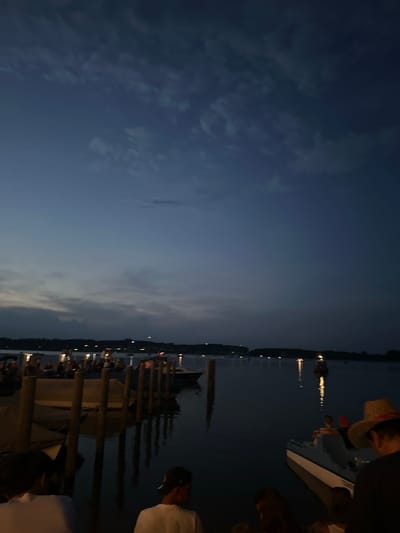 At dusk on a lake, several people are gathered on a dock. A few boats are moored nearby, and lights from a distant shore twinkle across the water. The sky is a deep blue, with wispy clouds tinged with grey. In the foreground, the backs of two individuals are visible, their faces obscured by the dim light. One of them wears a white t-shirt, while the other is seen from the side with a dark baseball cap. To the right, a person wearing a straw hat is seated in a small boat. The water reflects the dim lights and the dark forms of the dock pilings, creating a somewhat hazy and atmospheric scene. The location is the Chiemsee in Rimsting, Germany.