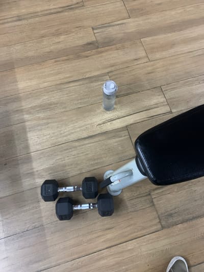 The image shows a weight bench in a room with wood-laminate flooring. The weight bench is a standard bench for weight lifting. There is a pair of 5kg dumbbells and another pair of 5kg dumbbells lying on the floor near the bench. A clear water bottle with a white lid lies on the floor at a distance from the weights. The coordinates -7.567, 110.757 place this location in a residential area in the city of Kartasura, Indonesia. A piece of footwear appears to be out of focus in the bottom right corner.