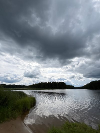 This image depicts a serene lake landscape under a dramatic, overcast sky. Dark, heavy clouds dominate the upper portion of the frame, with lighter, scattered clouds breaking through in places. The lake itself is calm, with gentle ripples reflecting the muted sky and surrounding dark green trees. The shoreline features lush green grass and a small sandy patch. The dense forest lines the far bank of the lake, creating a deep, shadowy contrast with the brighter parts of the sky. The scene suggests a natural, undeveloped environment, likely in a rural or wilderness area.