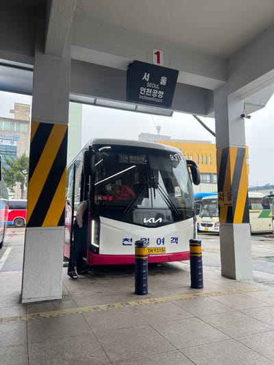 The image shows a bus stop in Geoje-si, South Korea. A white and pink bus, with the number 9315 on its windshield, is parked at the stop. The bus has "대한여객" (Daehan Passenger) written on its side. A sign above the bus indicates "1" and provides route information in Korean and English: "SEOUL Incheon Airport". Another sign on the bus reads "거제 통영동서울 (고현)" which translates to "Geoje Tongyeong-dong Seoul (Gohyeon)".
In the foreground, two dark blue bollards with yellow stripes are positioned near the bus. The bus stop area is paved with granite tiles, and a yellow line marks the edge of the bus lane. Yellow and black striped pillars are visible, typical of safety markings at transport hubs. In the background, other buses and buildings can be seen, suggesting a busy transportation center. The sky appears overcast, indicating potentially cloudy or rainy weather. The image is captured from a slightly low angle, looking up towards the bus and the bus shelter structure. There are no people clearly visible inside or around the bus, adding a sense of quietness to the scene. The overall impression is a functional transit stop on an ordinary day.