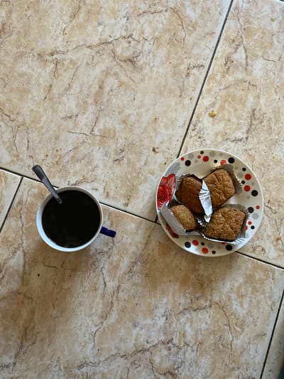 A top-down view of a white mug filled with black coffee, a spoon submerged within it, sits on a tiled surface. To the right, a white plate with red and black polka dots and a red floral design holds four pastries, each individually wrapped in silver foil. The tiled floor, composed of large, beige, marble-patterned tiles, shows faint streaks of dirt and small crumbs scattered across its surface. The scene appears to be indoors, possibly in a home setting within Baraki, Algeria, given the context of the request. There are no people visible in the image, and the objects are stationary.