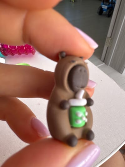 A brown, miniature capybara figure wearing a hooded costume is held by fingers with light pink nail polish. The capybara is holding a green cup with a white lid and straw, resembling a drink. The background is blurred but appears to be an indoor setting with a white surface, possibly a table, and hints of other objects and a doorway. The image is taken at close range, focusing on the capybara figure and the fingers. The location is in the city of Uyutnoye, Ukraine.