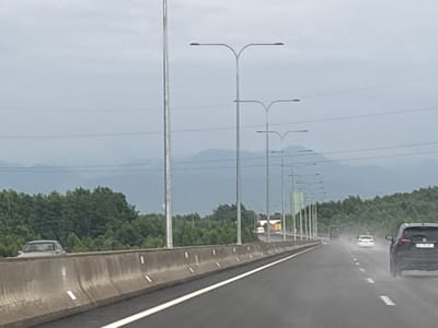 The image shows a highway in Shekvetili, Georgia, on a cloudy day. The road is wet, indicating it might be raining or has recently rained. Several cars are visible on the highway. In the foreground, a concrete barrier separates the lanes. Beyond the barrier, lush green trees line the road, and in the distance, mountains are visible under a hazy sky. Streetlights are mounted on poles along the highway. The overall scene depicts a typical highway environment in a natural setting.
