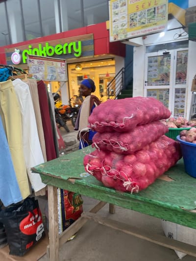 Three mesh bags of red onions sit on a green, weathered table next to a blue basket of smaller onions. In the background, a person wearing a blue turban stands near a store named "Frinkberry". Below the onions are rolls of fabric in shades of red, white, yellow, and blue. A black bag with red and white graphics is near the table's base.
