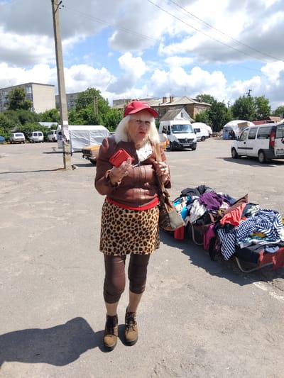 An elderly Caucasian woman wearing a red baseball cap, a brown leather jacket, a leopard print skirt, and brown leggings stands in a paved outdoor market area. She holds a red smartphone in her right hand and a dark brown purse on her left shoulder. In the background, several white vans and a tan car are parked. To the right of the woman, a large pile of folded and draped clothing is displayed on a cart. The environment appears to be a sunny day with scattered clouds in the sky. This scene is located in Chudniv, Ukraine.