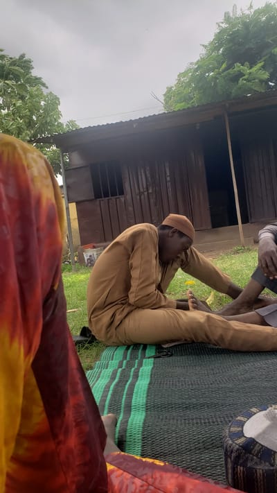 This media file captures an outdoor scene in Unguwar Shanu, Nigeria. The primary subject is a young man wearing a light brown traditional outfit and a matching cap. He is seated on a woven mat with green and black stripes, his legs bent in front of him. His gaze is directed downwards and to his right, with a thoughtful or perhaps slightly strained expression.
Another person's legs and feet are visible to his left, also seated. The background features a rustic wooden structure with a corrugated metal roof, suggesting a simple dwelling or workshop. Lush green foliage from trees flanks the scene on the left and top, and the ground is covered in grass, indicating a rural or semi-rural setting.
The sky is overcast, suggesting a cloudy or possibly rainy day, and the lighting is diffused, indicating it is likely daytime, perhaps late morning or afternoon. A colorful garment, predominantly red and yellow, is visible in the foreground, partially obscuring the view and framing the bottom of the image. The overall atmosphere feels relaxed and informal, with people gathered outdoors. No visible text is present in the image.