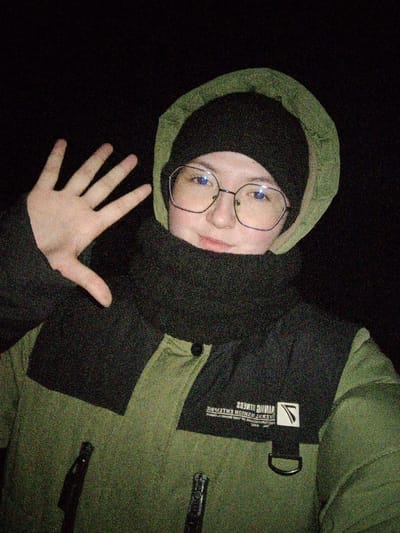 A nighttime, portrait-oriented image shows a single individual from the chest up, against a completely dark, featureless background, located in Старый Шагирт, Russia. The subject wears hexagonal eyeglasses, a black beanie, an olive-green hooded jacket with the hood up, and a black neck gaiter covering the lower face and neck. The individual's right hand is raised to the left of their head, palm forward with fingers splayed, in a gesture. A slight smile is present. The jacket features black shoulder and upper chest panels. Reversed white text reading "VIKING FITNESS" and a logo are visible on the right chest panel, along with a black D-ring attachment. The image exhibits significant digital noise, consistent with low-light conditions and flash illumination.