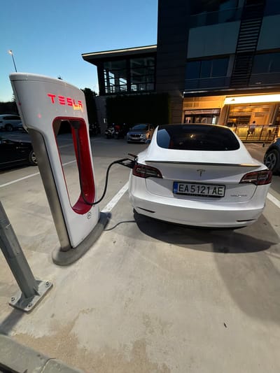 A white Tesla Model 3 is parked at a Tesla Supercharger station. The car is facing away from the camera, and a charging cable is plugged into its rear. The Tesla Supercharger station is white with red accents and features the Tesla logo. The scene is outdoors, with a concrete ground and a building in the background. The sky is dark blue, indicating it is either dawn or dusk. The license plate of the car is visible and reads "EA 5121 AE". The car is parked in a designated charging spot, marked by white lines on the concrete. Several other vehicles are parked in the vicinity, including a dark-colored car to the left and some motorcycles further in the background. The building in the background appears to be a modern structure with large windows and a lit entrance. The lighting suggests an ambient urban environment at twilight.