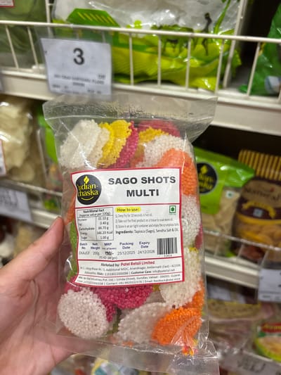A human left hand holds a transparent plastic pouch of "SAGO SHOTS MULTI" snack by the "Indian-chaska" brand. The product consists of multi-colored, raw tapioca (sago) pellets in white, yellow, pink, and orange. The label indicates a net weight of 200g and an MRP of ₹90/-. Nutritional facts, frying instructions, ingredients (Tapioca (Sago), Sendha Salt & Oil), and a 100% Veg symbol are displayed. The packing date is 25/12/2025, and the expiry date is 24/12/2026. A barcode with the number 32816 is present. The product is marketed by Patel Retail Limited, M-2, Udyog Bhavan No.: 5, Additional MIDC, Anandnagar, Ambernath (East) - 421506, Maharashtra, India, and manufactured by Ekai Industries Pvt. Ltd., Gundlav, Valsad, Gujarat. The background shows blurred white wire shelving typical of a supermarket or grocery store aisle, with other packaged food items in green and brown wrappers. A shelf price/item label marked "3" is visible above the held product.