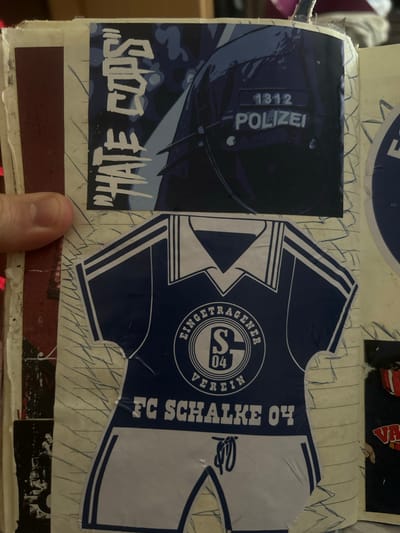 The image displays a collage of stickers pasted on what appears to be notebook pages. In the upper portion, a sticker features bold white lettering that reads "HATE COPS" against a dark blue background depicting a police helmet and uniform, with the word "POLIZEI" (German for Police) and the number "1312" visible. Below this, a sticker of a football jersey is prominent. The jersey is dark blue with white stripes on the sleeves and collar. A circular emblem on the chest contains the letters "S" and "04" within a shield shape, along with the text "EINGETRAGENER VEREIN" (registered association). The bottom of the jersey sticker reads "FC SCHALKE 04", indicating it represents the football club FC Schalke 04, which is based in the city of Gelsenkirchen, Germany. Other stickers, partially visible, include what appears to be a red and black design with text and a smaller black and white image of people. The background is lined paper, suggesting the collage is in a journal or scrapbook.