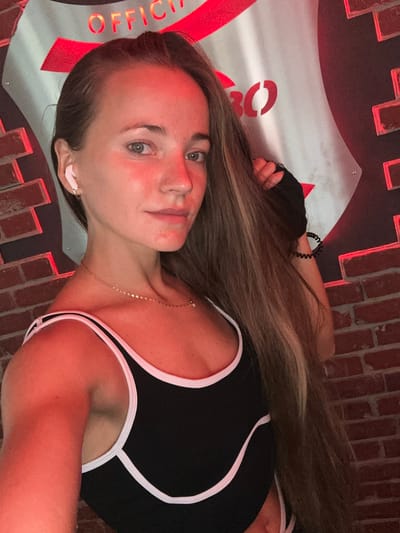The image shows a woman with blonde hair, wearing a black and white athletic top and white earbuds, standing in front of a brick wall and a metal sign. The sign has red lettering and appears to be a logo or crest. The lighting in the image is predominantly red, creating a dramatic effect. The woman is looking towards the camera, with her right hand touching her hair. The overall scene suggests an indoor setting, possibly a gym or a studio, given the athletic wear and the studio-like background elements. There are no visible indicators or clues that this location is in Wuppertal, Germany, or any specific city.