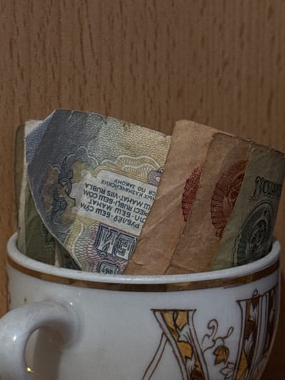A porcelain cup with a gold leaf pattern on the side is filled with several Soviet-era banknotes. The top note is faded and worn, with a visible denomination of "100". The note underneath has a prominent emblem of a hammer and sickle. The note below that has the text "РУБЛЁЎ БЕШ СЎМ ...". The cup is placed on a wooden surface. There are no people visible in the image. The location is not inferable from the image.