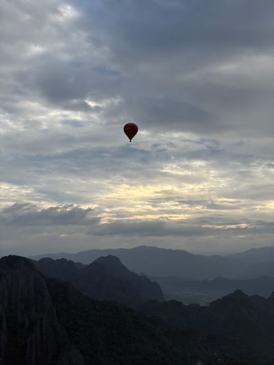 A hot air balloon is suspended in a cloudy sky above a mountain range. The balloon, colored red with yellow lettering, is small in the frame. The mountains below are dark and appear to be covered in dense vegetation. The sky is a mix of gray and white clouds, with some areas of lighter, possibly sunlit, sky. The overall scene suggests an early morning or late afternoon with the sun partially obscured by clouds. The location is Vang Vieng, Laos, known for its karst landscapes and outdoor activities such as hot air ballooning.