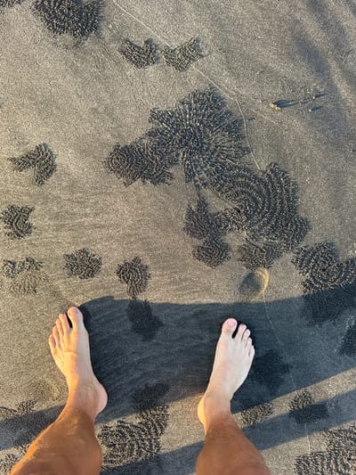 The image shows a pair of bare feet standing on a beach with dark, sandy ground. Circular patterns are visible in the sand, likely created by sand bubbler crabs. The lighting suggests it's either early morning or late afternoon. The location is presumed to be in the city of Selemadeg, Indonesia based on the prompt. No other people or objects are discernible. The action is simply standing on the sand. The environment is a beach.