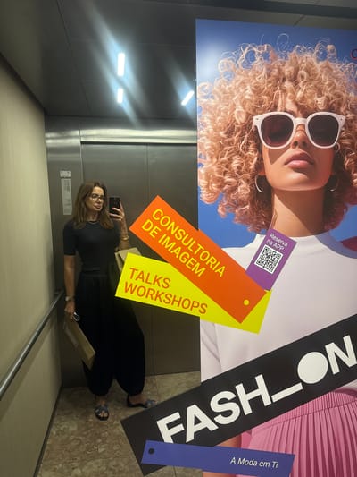 The image depicts a woman in an elevator, taking a selfie with her phone. She is holding a small box in her left hand, wearing a black outfit and blue sandals. She has brown hair and is wearing glasses. Behind her is an advertisement featuring a woman with blonde curly hair wearing sunglasses, with text in Portuguese. The text includes "CONSULTORIA DE IMAGEM," "TALKS WORKSHOPS," "Reserva na APP," and "FASH-ON A Moda em Ti." The elevator appears to be in the city of Matosinhos, Portugal. The elevator's interior is metallic, with visible lighting fixtures on the wall. The floor is tiled.