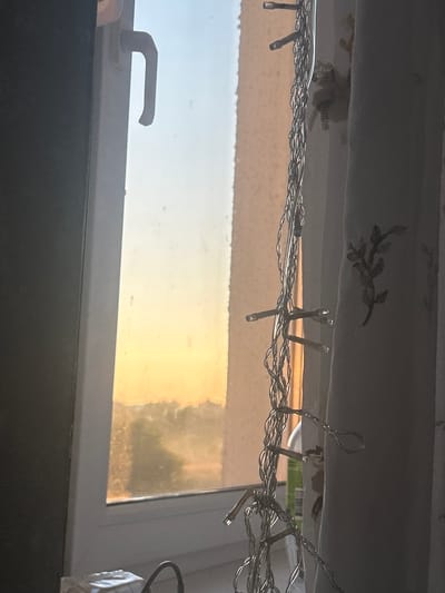 The image depicts an indoor scene through a window, with a view of a sunset. On the left, a portion of a dark, vertical surface is visible, possibly a wall or furniture. To the right of this, a white window frame is present, with a white handle in the upper left section of the frame. The window pane on the left shows a gradient of colors, transitioning from a pale blue sky at the top to a bright orange-yellow sunset glow at the bottom, with indistinct dark shapes in the distance suggesting trees or buildings.
Inside the room, a white curtain hangs on the right side of the frame, featuring a delicate embroidered pattern of a plant with leaves and branches in a darker thread. In the foreground, a string of silver fairy lights is draped vertically, with multiple small, cylindrical bulbs visible. These lights are intertwined with thin silver wire, creating a twisted effect. A white windowsill is below the window, on which a white tissue box and a black curved object, possibly a charging cable, are placed. The overall lighting suggests late afternoon or early evening. The location is specified as Яромінскі сельскі Савет, Belarus.