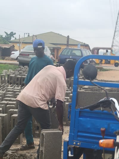 This outdoor scene captures two men engaged in manual labor involving concrete blocks. The primary subject in the foreground wears a light pink short-sleeved shirt and dark trousers, leaning over and appearing to handle a concrete block. Behind him, another man in a teal-colored short-sleeved shirt and a blue cap is also interacting with the blocks, which are stacked in multiple rows on the ground.
In the midground, a blue tricycle, possibly a cargo vehicle, is prominently featured, suggesting it is used for transporting the blocks. A black spherical object, likely a mirror or a light, is attached to the side of the tricycle.
The background shows a street environment with several vehicles. A blue car with a white license plate displaying "LAGOS STATE" and a faint "VW" (Volkswagen) logo is visible. Another lighter-colored sedan is parked further back. Buildings with pitched roofs line the street, and one building has the word "HALL" partially visible on its facade. Overhead power lines or cables crisscross against a bright, overcast sky.
The activity suggests the men are either loading, unloading, or stacking the concrete blocks, likely for a construction-related purpose. The time of day is daytime, under a hazy or overcast sky, providing soft, diffused light. The overall setting indicates a functional, possibly commercial or industrial area in Kwamba, Nigeria.