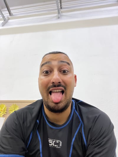The image shows a close-up of a man sticking his tongue out. He is wearing a black and blue long-sleeved shirt. There are two yellow paper flowers on a corkboard in the background, and a white wall behind him. Above the wall, there are metal beams. There is no visible indication of the location in Sevlievo, Bulgaria.
