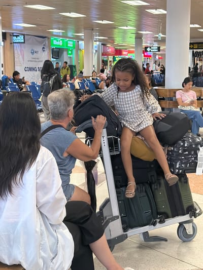 The image depicts a crowded indoor airport scene. A young girl with curly hair and a plaid dress is resting on a luggage cart piled high with suitcases. Her legs extend across the luggage. An older woman with gray hair sits nearby, appearing to steady the cart. Other people sit in rows of chairs in the background, some with luggage. A banner that reads "COMING SO" and mentions Chiang Mai International Airport is visible. An ATM and advertisements are also in the background. The image does not provide any details related to Kaungma, Myanmar.
