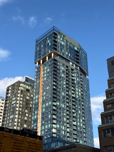 The image captures a low-angle view of a modern high-rise building with a grid-like facade of glass and metal, set against a clear blue sky with scattered clouds. The building's top portion appears to have a distinct cantilevered design. Other buildings are partially visible at the edges of the frame. The lighting suggests a sunny day. The location is not verifiable as Westmount, Canada without further information within the image.