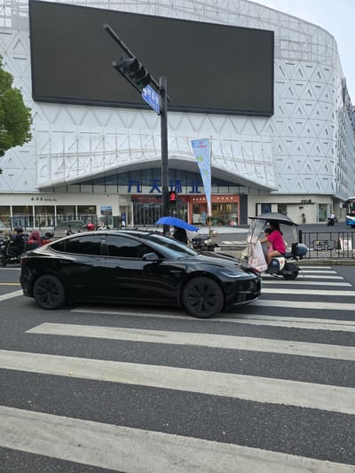 In Shangcheng District, China, a black Tesla is parked on a pedestrian crossing in front of a modern building with a distinctive geometric facade. The building's exterior features a large, dark digital screen and a white, textured surface composed of triangular patterns. A traffic light with a green signal is visible, indicating that vehicles may proceed.
Several people are present around a small, covered tricycle, with one person in a pink top and shorts leaning into the vehicle, possibly attending to a child lying on a blanket inside. The scene appears to be in daylight, and the weather is likely clear or partly cloudy.
Various signs with Chinese characters are visible on the building, including "米卡家 Mi Ka Jia" and "九天环北服装城 波函". A blue umbrella is also partially visible, suggesting it might be a sunny day or there's a possibility of rain. Several motorbikes are parked or moving in the background, typical of urban street scenes in China. The overall impression is a bustling urban environment with everyday activities taking place.