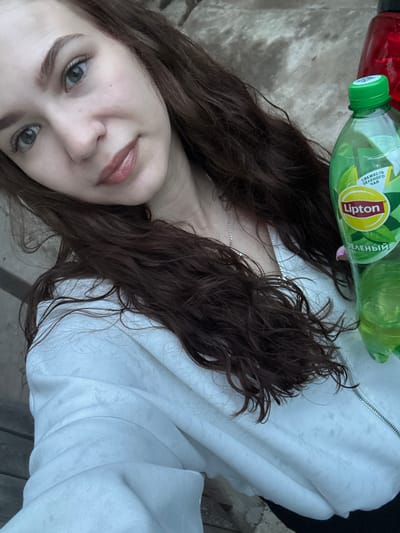 A woman is visible in a selfie-style image. She has brown hair, grey eyes, and light skin. She wears a white jacket with a zipper. Two plastic bottles, one with a green lid and the other with a red lid, are positioned near her neck. The green-lidded bottle has a "Lipton" label. The background appears to be an outdoor setting. Information on Nizhnyaya Gar', Russia is not discernible from the provided image.