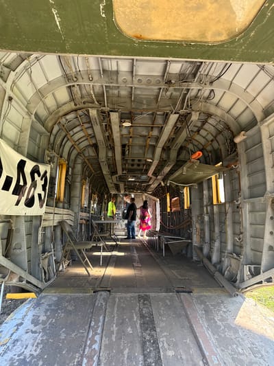 The interior of an aircraft fuselage is stripped bare, revealing the skeletal frame and curved walls. Light streams in from the open ends, highlighting the metal structure. Several people are visible towards the rear, seemingly exploring the gutted space. A white banner with large black letters is draped in the foreground. The interior exhibits a mix of metal textures, from smooth panels to rusted supports.